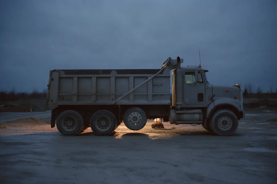 A tri-axle dump truck, three rear axles, side-on at first light